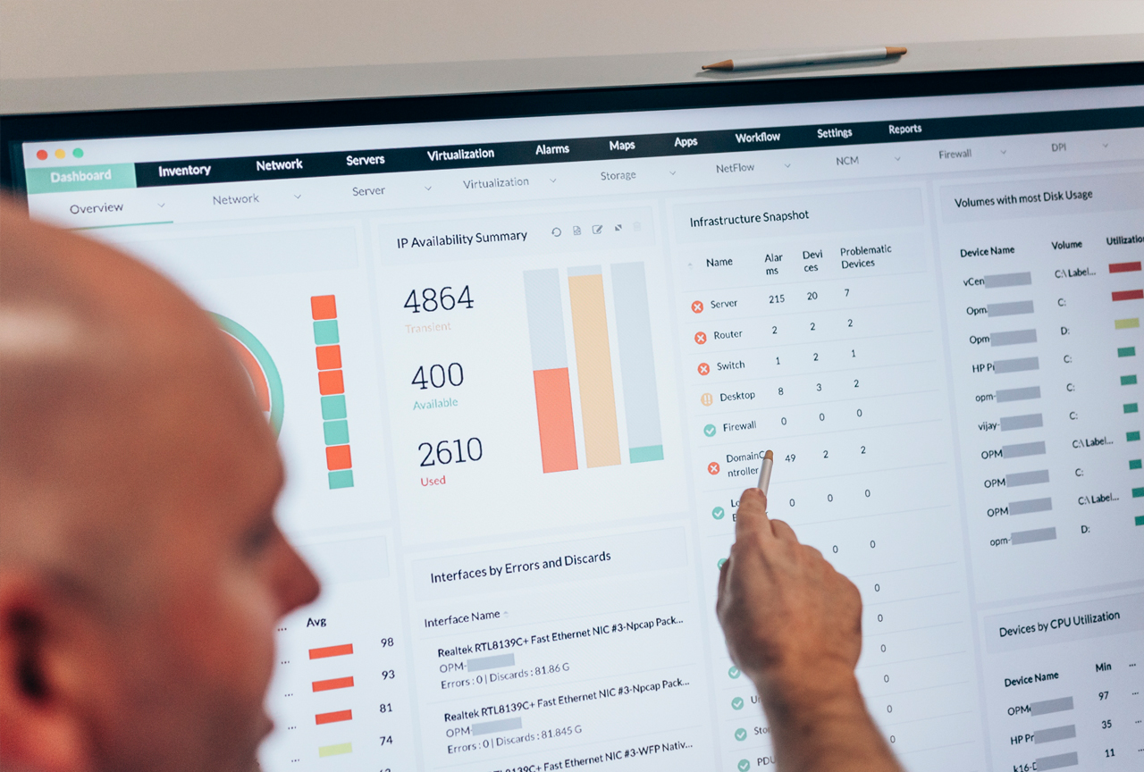 IT-Administrator zeigt mit einem Stift auf ein Monitoring-Dashboard mit Servern, Routern und Firewalls